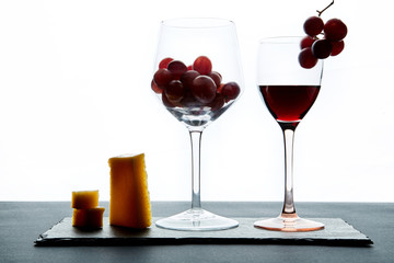 A few pieces of cheese and cherry tomatoes next to a glass of red wine filled with grapes on a white background