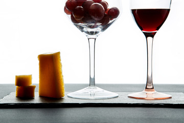 A few pieces of cheese and cherry tomatoes next to a glass of red wine filled with grapes on a white background