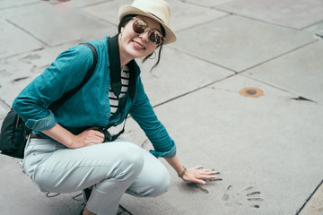 smiling asian woman traveler face camera looking joyful puts hand in handprint of famous star in chinese theater in hollywood los angeles california USA. young girl visit movie palace spring trip - obrazy, fototapety, plakaty