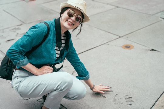 smiling asian woman traveler face camera looking joyful puts hand in handprint of famous star in chinese theater in hollywood los angeles california USA. young girl visit movie palace spring trip
