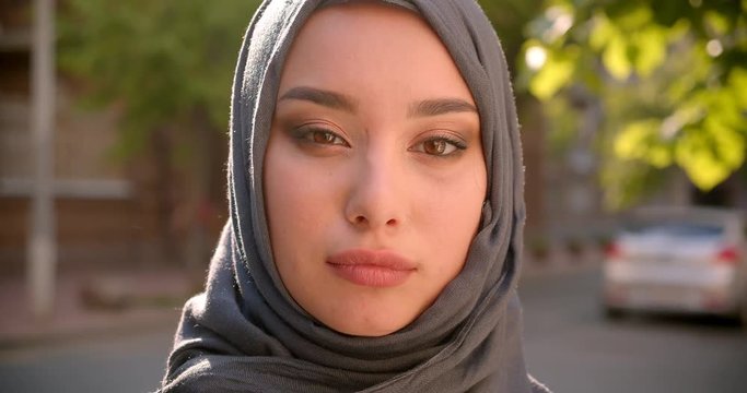 Portrait of muslim student in hijab watching calmly and modestly into camera standing on green city street.