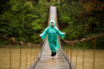 Traveller senior beautiful woman in blue rain jacket cross river by hinged bridge in forest, enjoying silence and harmony of nature