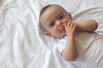Baby sucking his fingers. 6-8-month-old baby boy lying playfully in bed. Charming 6-7 month little baby in white bodysuit. Baby boy in white bedding. Copy space