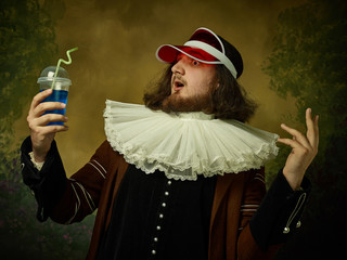 Young man as a medieval knight in red cap on dark studio background. Portrait of male model in retro costume. Holding a drink. Human emotions, comparison of eras and facial expressions concept.