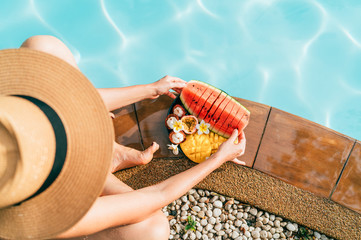 Caucasian Woman in straw hat sitting on swimming pool side near plate of tropical fruits- camera...