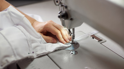 Female hands stitching white fabric on professional manufacturing machine at workplace. Seamstress hands holding lace textile for dress production. Blurred background. Close up view of sewing process.