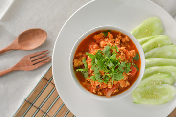  Top view delicious Northern Thai meat and tomato spicy dip in white bowl served with slice cucumber. Wooden spoon and fork.