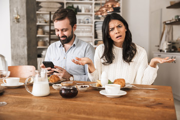 Portrait of european couple using mobile phone while sitting at table together during breakfast in kitchen at home