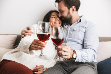 Image of romantic brunette couple drinking wine from glasses together while sitting on couch