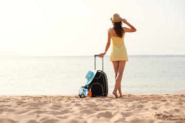 Young beautiful woman in yellow dress and hat with large suitcase on tropical beach. Girl look on sea