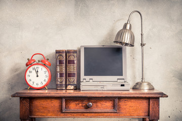 Vintage alarm clock, antique old books, outdated portable notebook computer from 90s and desk lamp on the oak wooden table front loft concrete wall background. Retro style filtered photo