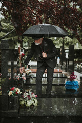 Elegant sad elderly man standing on the rain with umbrella and grieves at the grave of a loved person