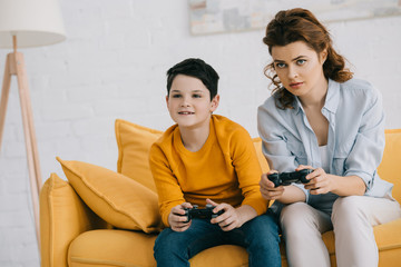 Fototapeta premium focused mother and son playing video game with joysticks while sitting on sofa at home