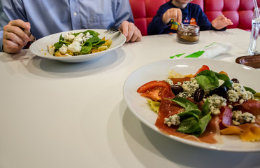 Delicious Italian pasta with vegetables and herbs in a plate on a white table in a restaurant. In the background a man with a child is eating at the table