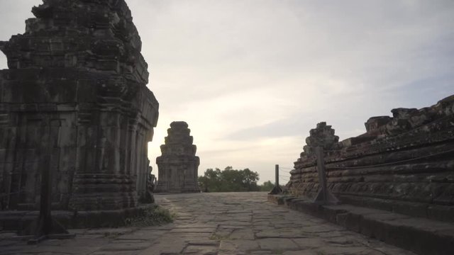 Slowly walking across the top of a Khmer temple at sunset; setting sun revealed from behind temple.