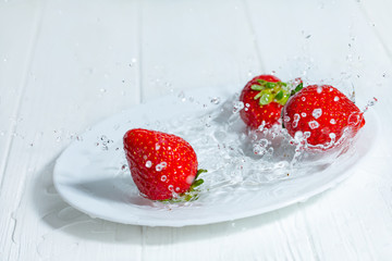 Fresh strawberries fall on a white plate. Splashes of water around the berries. White wooden table.