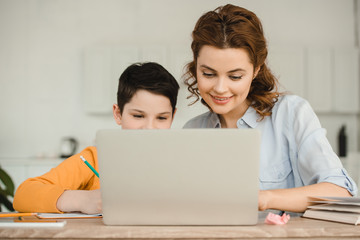 happy mother with cute son using laptop together while doing schoolwork at home - obrazy, fototapety, plakaty