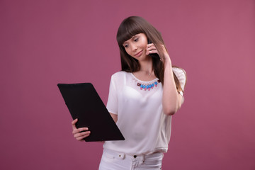 Image of young business woman standing over pink background looking aside chatting by phone holding folder.