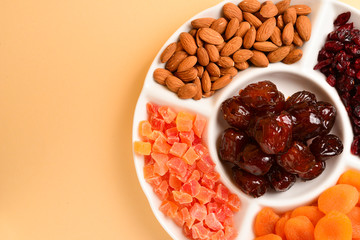 Mix of dried fruits and nuts on a white plate. Apricot, almond, raisin, dates fruit. On a brown background. Space for text or design.