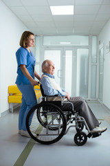Fototapeta na wymiar friendly nurse pushing senior patient in wheelchair at hospital corridor