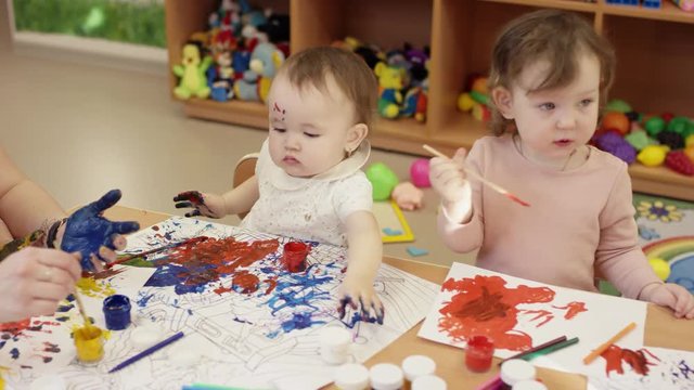 children sit at the table and draw, a little girl picks up a white jar of paint