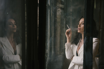 Fototapeta premium Young sexy brunette woman in suit smoking a cigarette by the window indoors