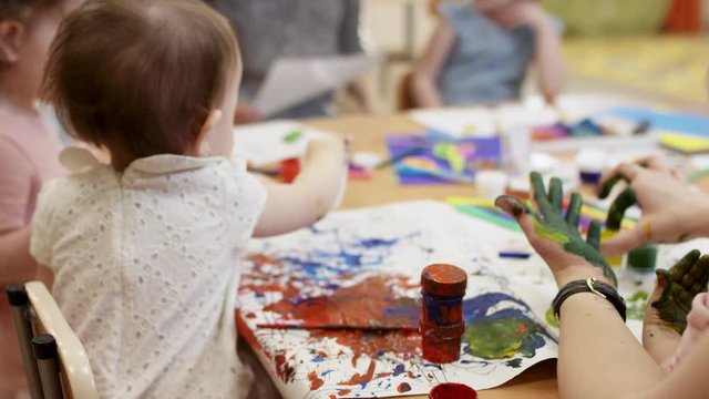 children sit at the table and draw with paints, on the right you can see the hands of the kindergarten teacher, they are green