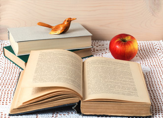 Still life. On a table covered with a white lace tablecloth are books. One textbook is open for reading. In the background is a red apple and a wooden bird.