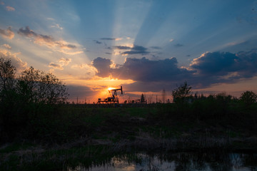 Equipment for oil and gas production works at sunset in the rays and glare of the sun next to the river. Oil well at sunset. Oil production with rocker at sunset. Rocking oil