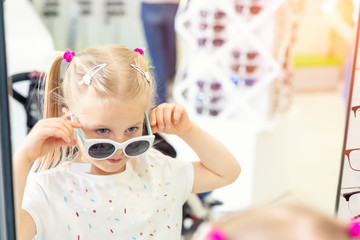 Cute little young caucasian blond girl trying on and choosing sunglasses in front of mirror at optic eyewear store. Adorable schoolgirl child having fun bying sun uv protection glasses at shop