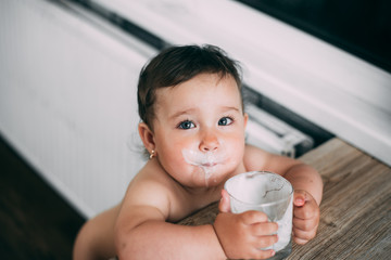 A little girl in the kitchen drinking milk or yogurt from a glass is very appetizing and greedy all face dirty