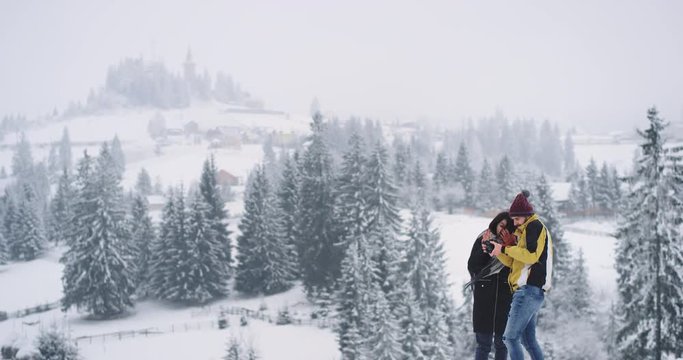 Young attractive guy and lady standing on the top of field background amazing snowy forest , they looking at the pictures of they take before