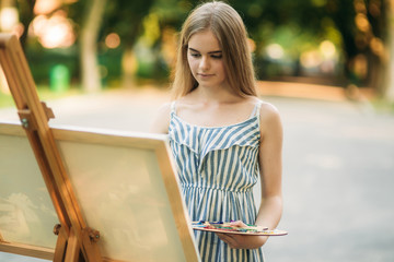 Beautiful girl draws a picture in the park using a palette with paints and a spatula. Easel and canvas with a picture