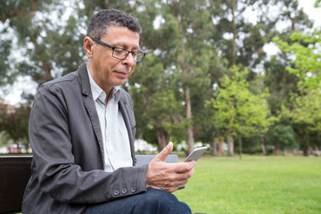 Content man using smartphone and sitting on bench in park. Guy wearing casual clothes and texting on gadget with green lawn and trees in background. Communication and nature concept.