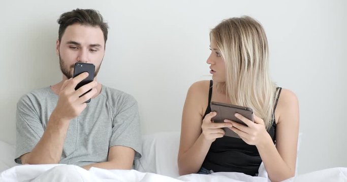 Couple in bed using mobile phone and tablet together, woman dislike her man of watching too much phone. Caucasian man and woman.