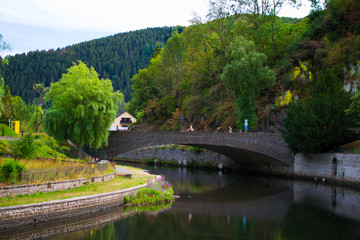 Old stone bridge over Sure river with a walkway through the riverside and mountain and trees at the background. Esch Sur Sure, Luxembourg
