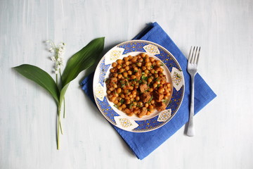 chickpea with meat made in red sauce served in white and blue plate with fork and lily of the valley flower
