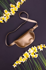 Bright summer fashion ladies accessories. Stylish gold leather handbag or fanny pack on a grey background with narcissus or daffodil flowers in corner. Top View. Flat Lay. Mock up.