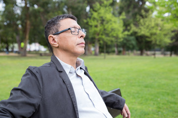 Relaxed middle-aged man sitting on bench in park. Guy wearing casual clothes and keeping his eyes closed with green lawn and trees in background. Relaxation and nature concept.