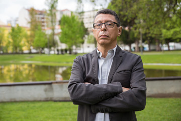 Serious middle-aged man posing at camera and standing in park. Guy standing with his arms crossed,...