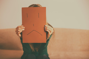 Depressed woman sitting alone at sofa and holding paper with sad face.Toned image.