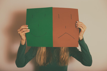 Woman with happy and sad face on paper standing  in front of wall.Toned image.