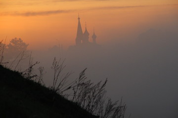 Obraz premium Silhouettes of church, trees, hill grass in misty morning 
