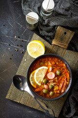Traditional Russian soup Solyanka with meat, sausages, vegetables, capers, pickles and olives with lemon on black background. Rustic style. Top view flat lay background.