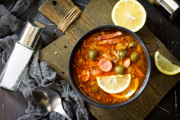 Traditional Russian soup Solyanka with meat, sausages, vegetables, capers, pickles and olives with lemon on black background. Rustic style. Top view flat lay background.