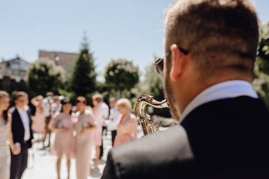Saxophonist in Black Jacket and Glasses on Wedding Ceremony with Soft Focus. Talanted Male Musician Play on Jazz Instrument for Guests on Romantic Happy Celebration Outdoor Background