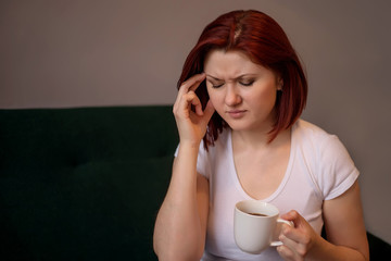 Exhaustion, burning out, migraine or headache concept: woman with bob haircut sits on sofa with white cup of coffee touching or massaging her temple