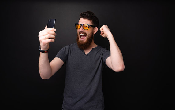 Photo of excited bearded man holding mobile, scraming and celebraing with rised hands