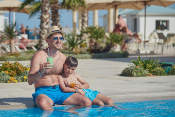 Dad and son enjoying their summer holidays. They are holding their cocktails in the hands, while sitting near the hotel’s swimming pool.