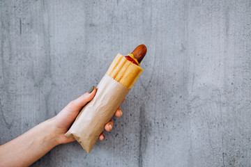 French hot dog in a bacon with fried sausage in a woman's hand against the wall